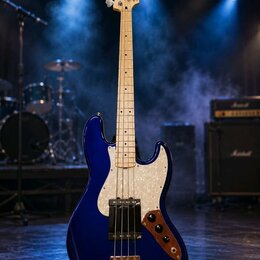 Modrá elektrická basová kytara s bílým hmatníkem a bílým perleťovým pickguardem. Na hlavě je nápis Fender Jazz Bass. Nástroj je umístěn na stojanu v…