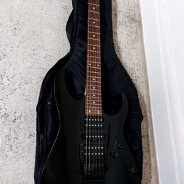 Černá elektrická kytara s tmavým hmatníkem, umístěná v černém poloměkém pouzdře. Na hlavě kytary je bílý nápis Ibanez. Nástroj má dva humbuckery, ovl…