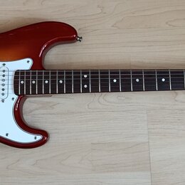 Elektrická kytara typu Stratocaster s tělem v červeně-žlutém sunburst odstínu a bílým pickguardem. Na hlavě hmatníku je vidět nápis Squier STRATOCAST…