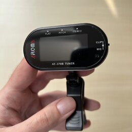 Černý digitální ladicí přístroj s displejem a připevněným klipem. Na levé straně je červené logo AROMA a v dolní části nápis AT-370B TUNER. Horní hra…