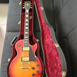 Elektrická kytara typu Les Paul s červeno-žlutým sunburst povrchem v černém tvrdém kufru s červeným plyšovým vnitřkem. Na hlavě hmatníku je nápis Gib…