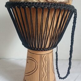 Djembe s tělem z vyřezávaného světlého dřeva a napnutou kůží v horní části. Nástroj je opleten černými provázky, které drží membránu. Spodní část kor…