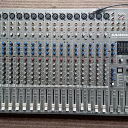 Šedý mixovací pult s nápisy SAMSON, L2000 LIVE MIXING CONSOLE a 24 DSP USB. Zařízení obsahuje řadu vstupních kanálů s modrými a červenými knoflíky, p…
