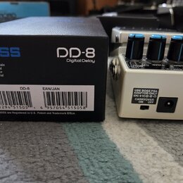 Na fotografii je kytarový efekt Boss DD-8 Digital Delay v bílém provedení s modrými ovládacími knoflíky. Vedle pedálu je umístěna originální černá kr…