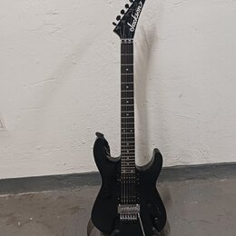 Černá elektrická kytara s špičatou hlavou, na které je bílý nápis Jackson. Nástroj má tmavý hmatník a stříbrný kobylku typu Floyd Rose. Kytara je umí…