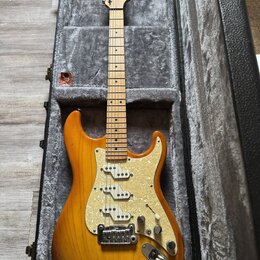 Elektrická kytara s přechodovým žlutě-hnědým lakem v černém tvrdém kufru. Nástroj má bílý pickguard s perleťovým vzorem, tři snímače a javorový hmatn…