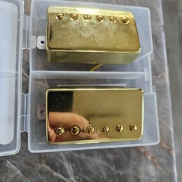 Dvě elektrické kytarové snímače typu humbucker v plastovém průhledném pouzdře. Oba mají zlatý kovový kryt a šest viditelných šroubů. Povrch horního s…