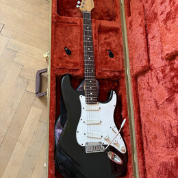 Černá elektrická kytara typu Stratocaster v hnědém tvrdém kufru s červeným plyšovým vnitřkem. Na hlavě hmatníku je vidět nápis Fender. Kytara má bílý…