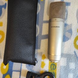 Kondenzátorový mikrofon stříbrné barvy s mřížkou v horní části a nápisem M-AUDIO na těle. Vedle mikrofonu je černý kožený obal se zipem a černý plast…