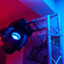 Na kovovém hliníkovém trussu jsou upevněna čtyři světla. V popředí je velký černý pohyblivý hlavový reflektor svítící modře, za ním následují tři men…