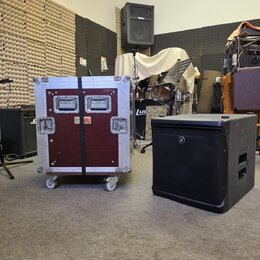 Na fotografii je vidět transportní flightcase v červené barvě s hliníkovými hranami a koly, vedle kterého stojí černý subwoofer. Na flightcasu jsou u…