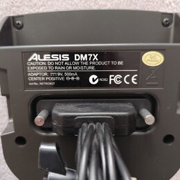 Detail zadní strany černého elektronického zařízení s několika připojenými kabely. Na stříbrném štítku je uveden název Alesis DM7X a varovný text CAU…