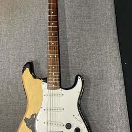 Elektrická kytara typu Stratocaster s černým lakem, který je výrazně odloupaný a odhaluje světlé dřevo korpusu. Na hlavě hmatníku je nápis Fender. Ky…