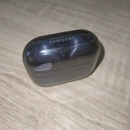 Černý nabíjecí box s průhledným víkem, uvnitř kterého jsou umístěna dvě bezdrátová sluchátka. Na horní části víka je bílý nápis SAMSUNG. Povrch průhl…