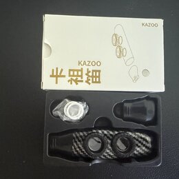 Bílá krabička s nápisy KAZOO a čínskými znaky 卡祖笛 obsahuje černý kazoo s povrchem v optice karbonu. V plastovém vložném lóži je vedle nástroje umístě…