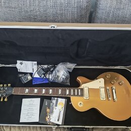 V otevřeném kufru leží elektrická kytara typu Les Paul v zlatavé barvě s krémovým pickguardem. Na hlavě kytary je nápis Gibson a modelové označení De…