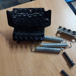 Černý kovový kobylník s nápisem Licensed under Floyd Rose Pats. Vedle něj leží tři kovové pružiny, jedna kovová opěrka a dva černé šrouby. Všechny dí…