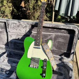 Zelená elektrická kytara typu T-style s bílým pickguardem v otevřeném hrdelním kufru. Na hlavě hmatníku je vidět nápis Fender. Kytara má připevněný v…
