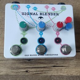 Bílý efekt pedál s nápisem SIGNAL BLENDER a v dolní části OLD BLOOD NOISE ENDEAVORS. Zařízení má tři stoupací spínače s barevnými LED diodami (zelená…