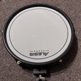 Kruhová elektronická bubenová pad v černé barvě s bílou úderovou plochou. Na spodní části plochy je černý nápis ALESIS DRUMS. Pad má černý obrubník a…