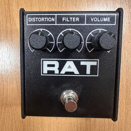 Černý kytarový efekt pedál s nápisem RAT v bílém rámečku. Horní část obsahuje tři černé otočné knoflíky s popisky DISTORTION, FILTER a VOLUME. V doln…
