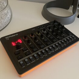 Černý sekvenční syntetizátor s oranžovými bočními panely. Na zařízení je bílý nápis Roland a model T-8, červený digitální displej zobrazující znaki „…