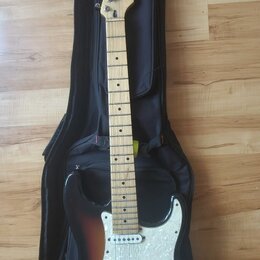 Elektrická kytara s tmelem v sunburst provedení a bílým pickguardem, položená na černém obalu. Na hlavě hmatníku je vidět logo a nápis Stagg. Nástroj…