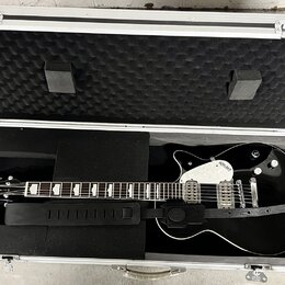 Otevřený hliníkový kufr s černou pěnovou výplní, ve kterém leží černá elektrická kytara a černý popruh. Na hlavě kytary je nápis GRETSCH. Tělo kytary…