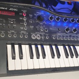 Černý syntetizátor s klaviaturou a ovládacím panelem s knoflíky a tlačítky. Na horní části je bílý nápis Roland JD-Xi a červeně svítící displej s tex…