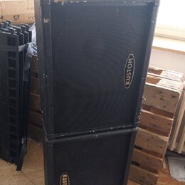 Dvě černé basové bedny s kovovými rohy, naskládané na sebe. Obě mají na přední mřížce oválný nápis KUSTOM. Povrch horní bedny vykazuje v horní části…