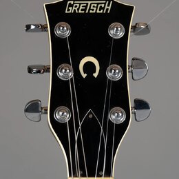 Detail hlavy elektrické kytary značky GRETSCH v černé barvě s bílým lemováním. Uprostřed hlavy je umístěno bílé písmeno C. Jsou vidět šest chromovaný…