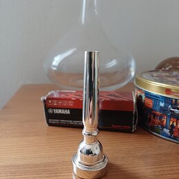 Stříbrný, kovový mute (tlumík) s válcovým tvarem a rozšířenou základnou. Na spodní části je vyryt nápis "JEAN-PIERRE". V pozadí je vidět červená krab…