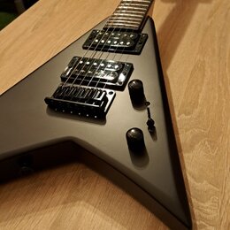 Matně černá elektrická kytara s tělem ve tvaru V položená na dřevěném stole. Nástroj má dva humbuckery, jeden potenciometr hlasitosti a jeden tónový…
