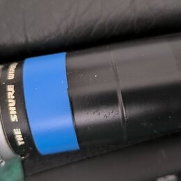 Detailní pohled na horní část černého bezdrátového mikrofonu s kovovou síťkou. Na těle je modrý ozdobný kroužek a nápis THE SHURE WIRELESS. Jsou vidě…