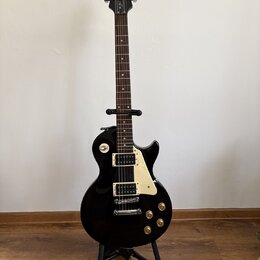Černá elektrická kytara typu Les Paul značky Epiphone, umístěná na černém stojanu. Na hlavě hmatníku je bílý nápis Epiphone a vpravo pod ním kurzivní…