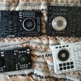 Fotografie zobrazuje čtyři DJ kontrolery položené na vzorité přikrývce. V horní levé části je černý kontroler s nápisy serato a Numark. V horní pravé…