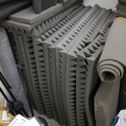 Svrhovatě naskládané šedé akustické pěny s pyramidovitým povrchem. Vedle nich je srolovaný šedý zvukový izolant a další kusy pěnového materiálu. V po…