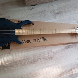 Tmavě modrá elektrická basová kytara položená na kartonovém obalu. Na obalu je svisle napsán text Marcus Miller. Hmatník je zC světlého dřeva a hlava…