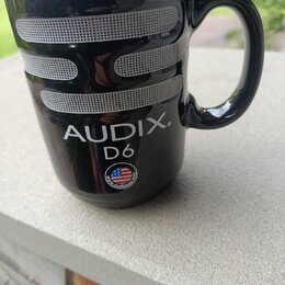 Černý keramický hrnek ve tvaru mikrofonu s vymalovanou stříbrnou mřížkou. Na povrchu je bílý nápis AUDIX. D6 a malá kulatá nálepka s vlajkou USA a ná…