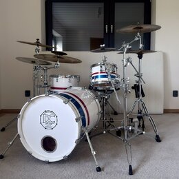 Bílá bicí souprava s modrým a červeným pruhem. Na přední straně basového bubnu je logo s nápisem SINCE 2004, DC DRUMS a QUALITY INSTRUMENTS. Souprava…