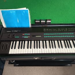 Černý digitální syntezátor s klávesnicí, na kterém je nápis YAMAHA DX7. Nástroj je umístěn v otevřeném transportním kufru s zeleným vnitřkem, ve kter…