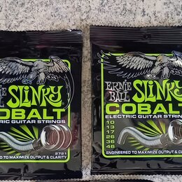 Dvě uzavřená balení kytarových strun v černých obalech s limegreenovými prvky. Na obou je nápis ERNIE BALL SLINKY COBALT ELECTRIC GUITAR STRINGS a te…