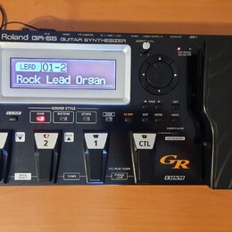 Černý kytarový syntetizátor s nápisem Roland GR-55 GUITAR SYNTHESIZER. Zařízení má digitální displej, na kterém je text LEAD 01-2 Rock Lead Organ, ně…