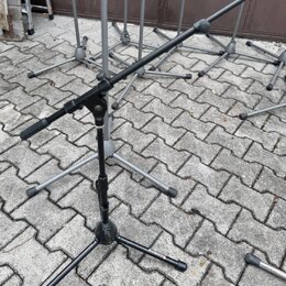 Fotografie zobrazuje několik černých kovových stativů na mikrofony s trojnožkou, umístěných na dlažbové ploše. Na jedné z nožek předního stativu je b…