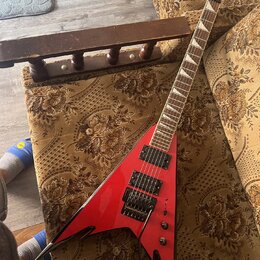 Červená elektrická kytara s tělem ve tvaru V a černými hranami. Na hlavě hmatníku je bílé logo Jackson. Kytara má dva černý humbuckery, černý most s…