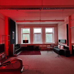 Fotografie zachycuje interiér místnosti s červeným osvětlením. V prostoru je umístěn černý digitální klavír s bílými klávesami a černá pohovka. V lev…
