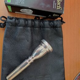 Stříbrný kovový moutur položený na černém koženém vakové pouzdře. Vedle nich je černá krabička s nápisy Bach Genuine Mouthpiece a LEAD FREE. Na moutu…