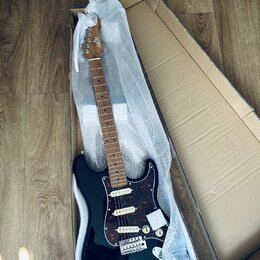 Černá elektrická kytara s tmavě červeným šarlatanem (pickguard) v kartonové krabici s ochrannou bublinkovou fólií. Na hlavě hmatníku je bílý nápis "S…