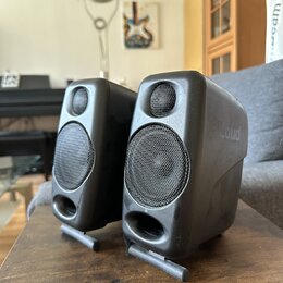 Dva černé plastové reproduktory s mřížkami přes membrány, umístěné na dřevěném stole. Na pravém reproduktoru je vylisovaný nápis Loud. Oba přístroje…