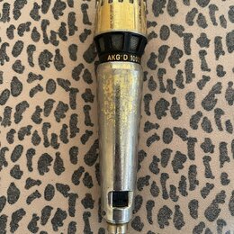 Zlatý dynamický mikrofon s mřížkou v horní části. Na těle je černý pruh s žlutým nápisem AKG D 1000. Povrch mikrofonu vykazuje výrazné opotřebení, šk…